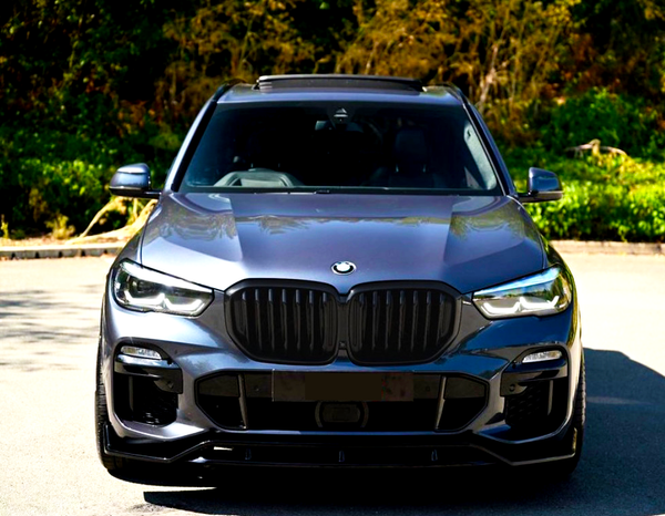 A dark-colored car is parked on a sunlit road, surrounded by lush green foliage. The car's front features a distinctive grille and sleek headlights.