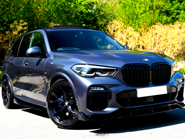 A dark-colored SUV is parked on a driveway, facing forward. It features a sleek design with large alloy wheels, positioned against a backdrop of lush greenery and sunlight.