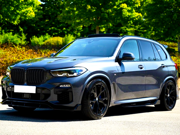 A parked gray SUV with tinted windows and black wheels is in a sunlit parking lot, surrounded by green trees and low shrubs.