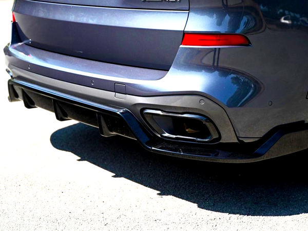 A dark metallic car's rear end, featuring dual exhaust tips and a diffuser, is parked on a sunlit asphalt surface. The vehicle's body reflects nearby buildings and trees.