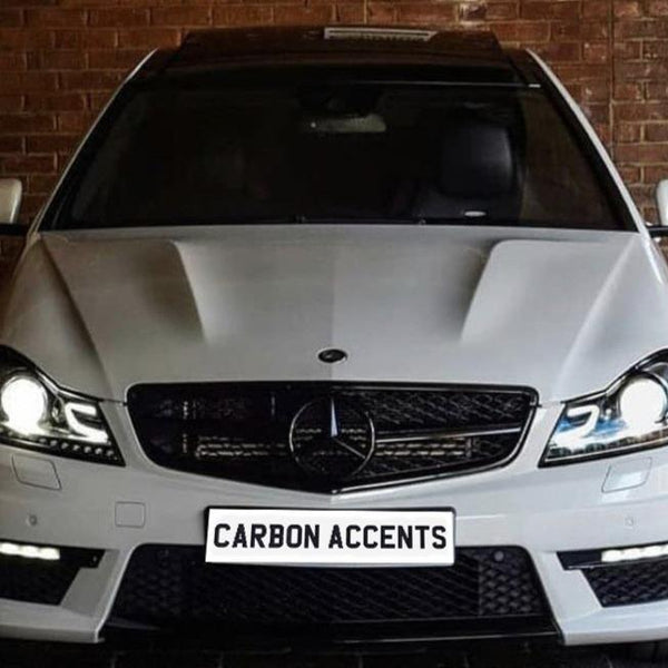 A white Mercedes-Benz car with illuminated headlights is parked in front of a brick wall. The license plate displays the text "CARBON ACCENTS."