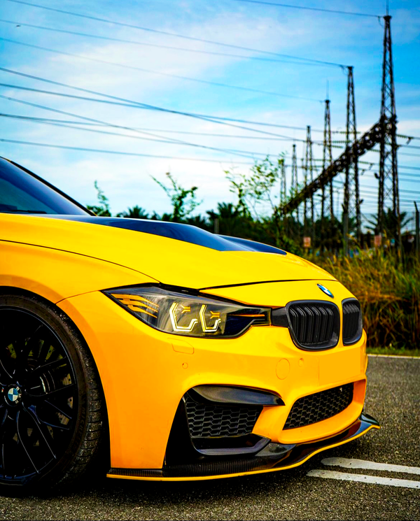 A yellow sports car, featuring sleek black accents and shining headlights, is parked on a road. Its background includes tall electrical towers and a clear blue sky.