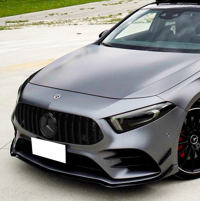 A sleek, silver car is parked on a concrete surface beside a grassy area. It features a prominent grille and glossy black wheels with red brake calipers.