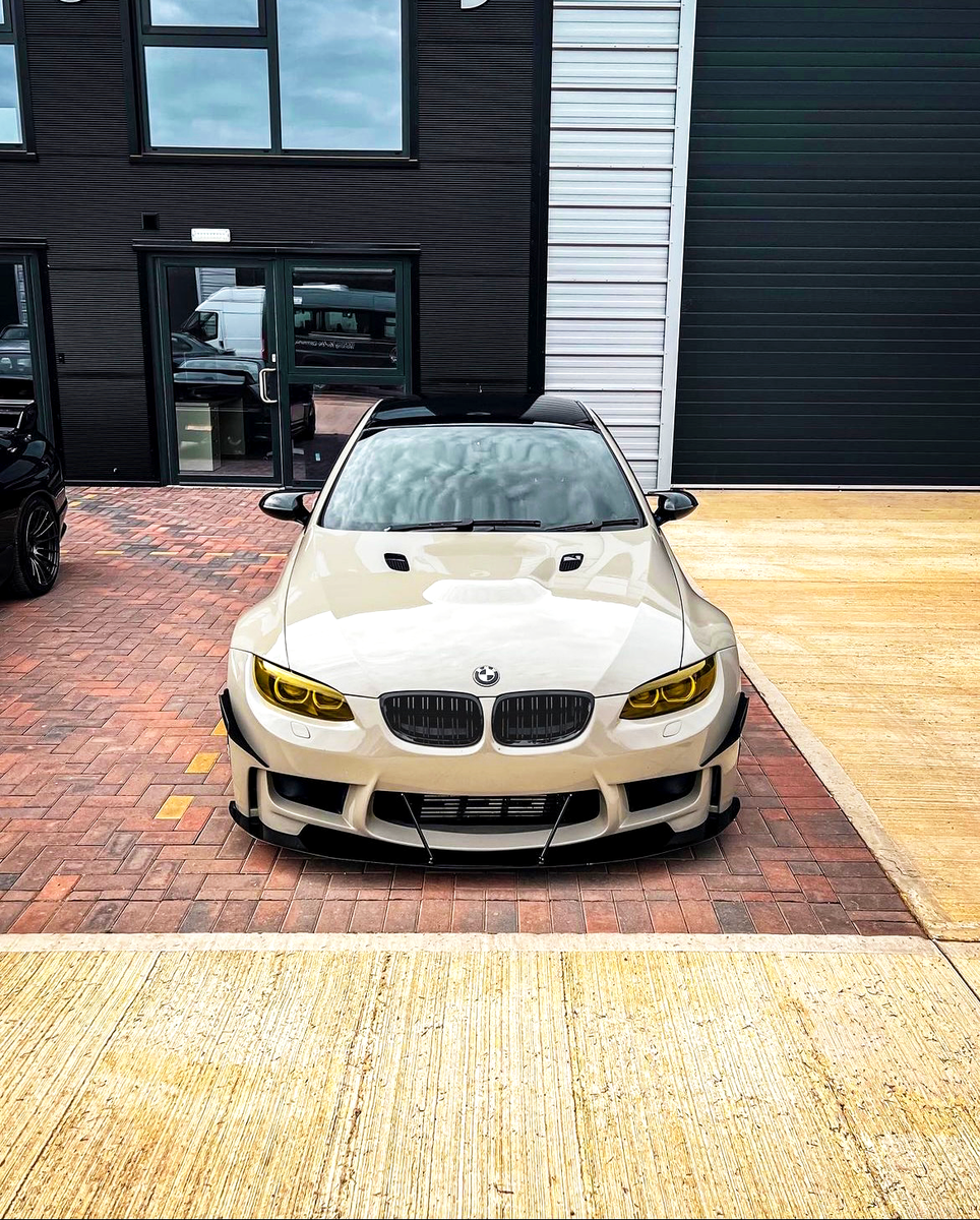 A white car with yellow-tinted headlights is parked on a brick driveway in front of a modern building with large windows and a garage door.
