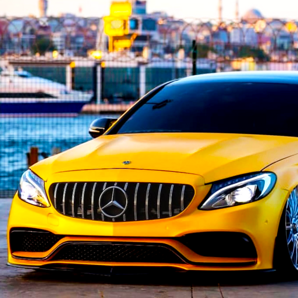 A yellow Mercedes-Benz car is parked near a waterfront with a city skyline in the background, including distant buildings and a boat on the water.