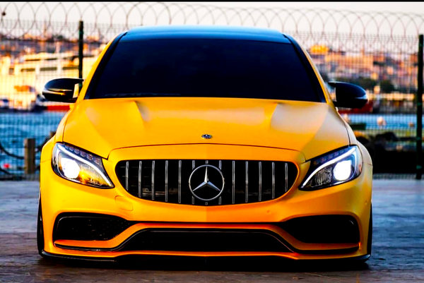 Yellow Mercedes-Benz car is parked, facing forward with headlights on, in front of a wire fence. The background includes a blurred water scene and distant buildings.