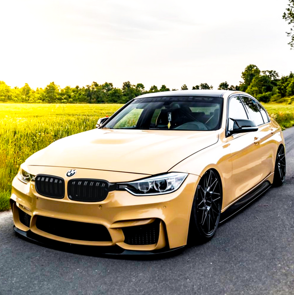 A beige BMW sedan parked on a rural road, surrounded by green fields and trees under a bright sky. The car features black rims and a sleek body.
