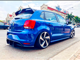 A blue Volkswagen hatchback with aftermarket rims is parked on the street. It features tinted windows, and a rear spoiler, with a building and palm trees in the background. Text: 