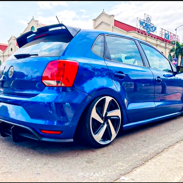 A blue Volkswagen hatchback with aftermarket rims is parked on the street. It features tinted windows, and a rear spoiler, with a building and palm trees in the background. Text: "@v_w_p_olo."