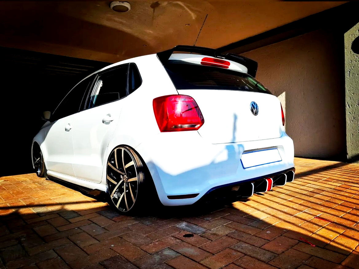 A white car parked on a brick driveway under a shaded carport. The car features large, black alloy wheels and a distinctive rear bumper. Shadows create a contrasting pattern on the ground.