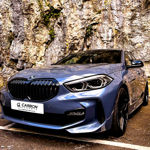 A blue car is parked in front of a rocky wall, showcasing a sleek design. The front license plate reads, "CARBON ACCENTS www.carbonaccents.co.uk."