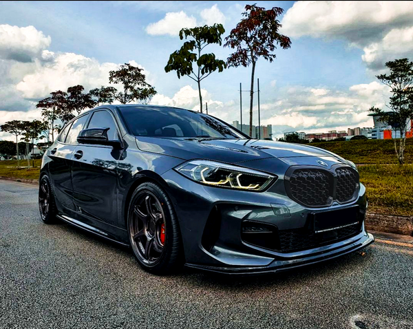 A sleek, dark gray car is parked on a road, surrounded by sparse trees and buildings under a partly cloudy sky.