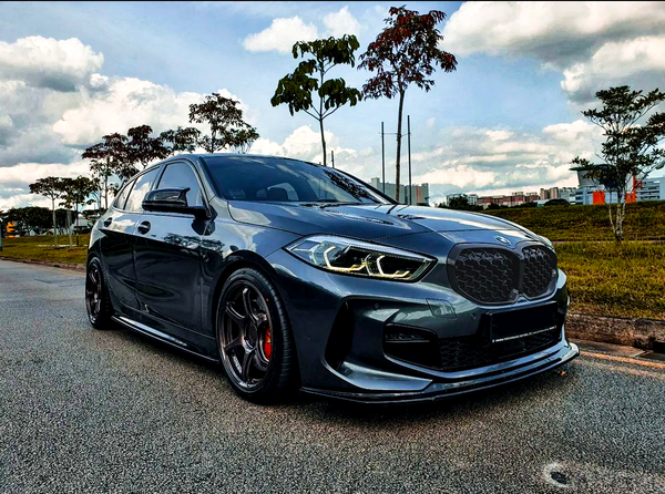 A sleek, dark gray car is parked on a road beside grassy areas and trees under a partly cloudy sky, with distant buildings visible.