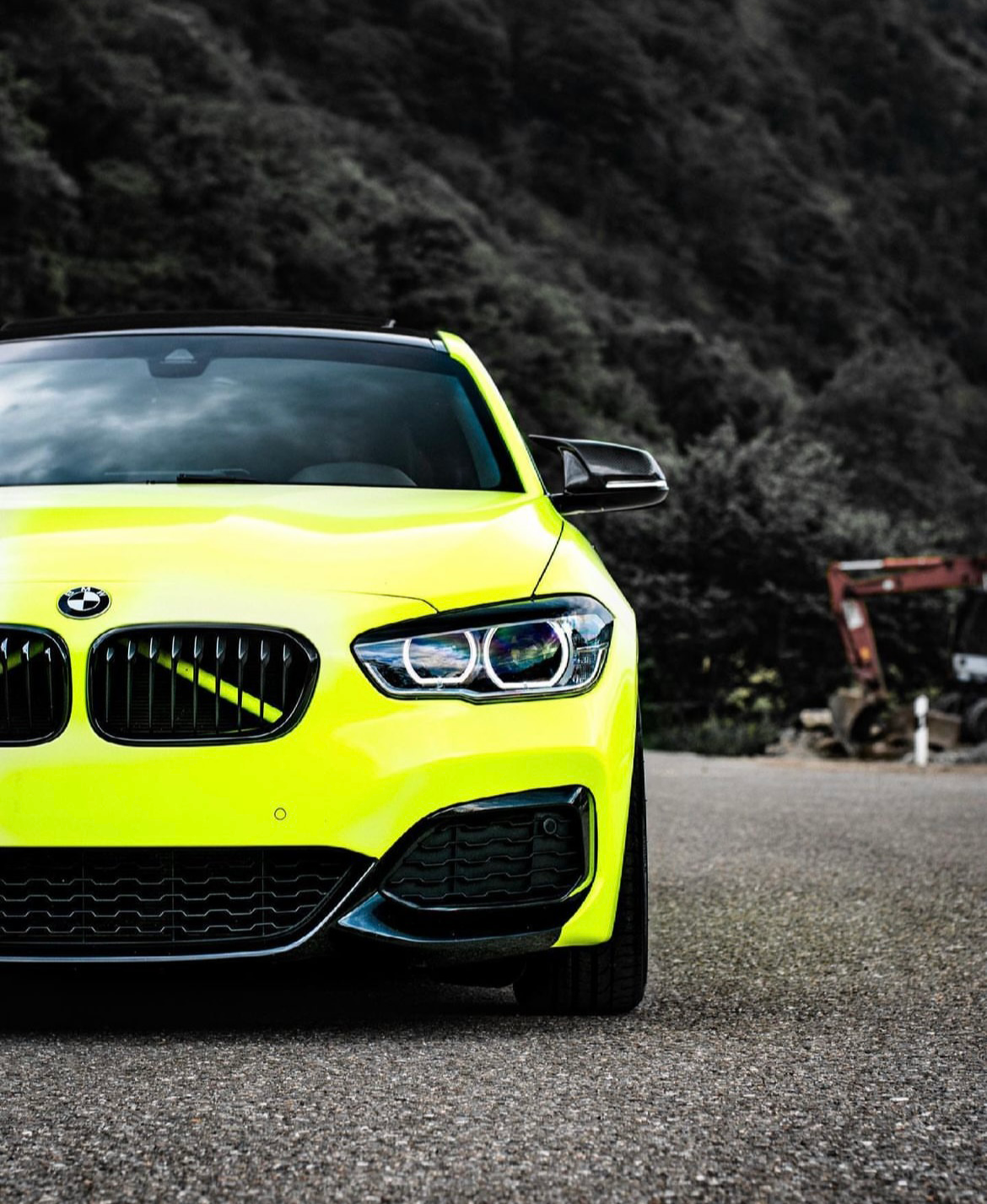 A bright yellow BMW car stands parked on a road, its sleek design contrasting with a forested hillside in the background. The environment appears rural with scattered machinery visible.