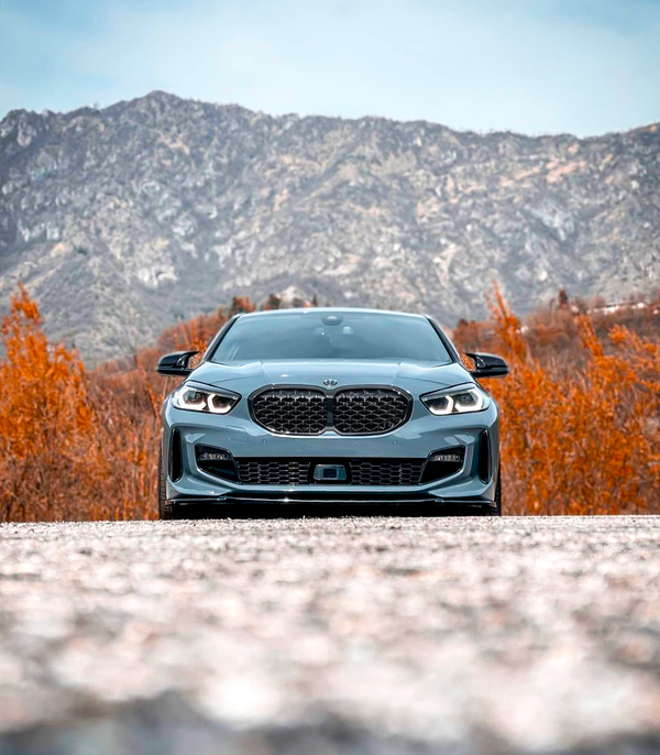 A sleek gray car is parked on a gravel road, with vibrant orange foliage and towering mountains in the background, under a clear blue sky.