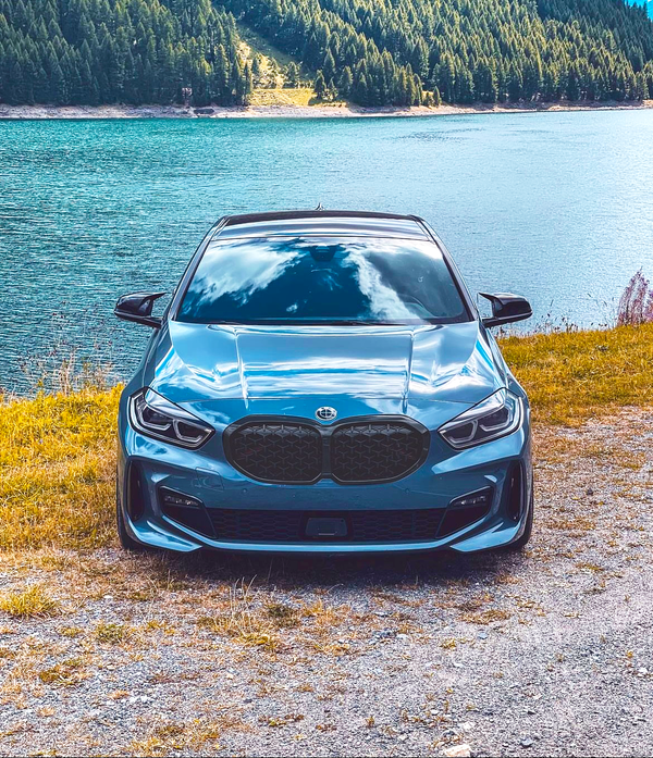 A blue car is parked on gravel near a lake, reflecting clouds. Behind, dense green forest rises over hillsides, contributing to a serene, natural landscape.