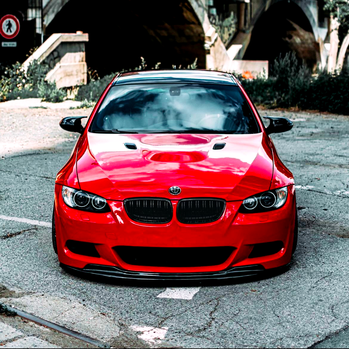 A shiny red sports car with a sleek design is parked on a cracked pavement, under a bridge with greenery surrounding it. A circular road sign is visible in the background.