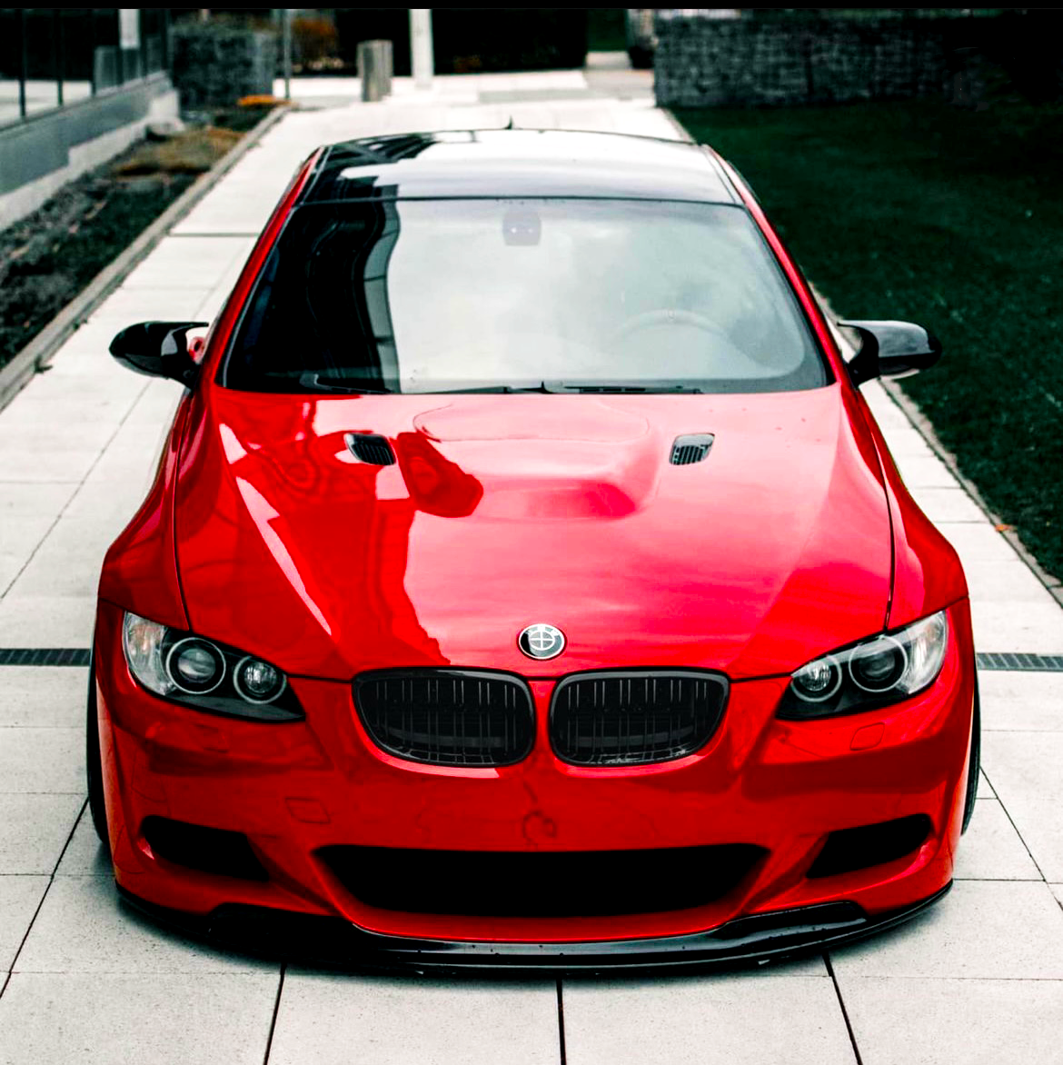 A red sports car, parked on a tiled pathway, features a sleek design with aerodynamic curves and a shiny finish. Dense urban surroundings include a grassy area and modern buildings.