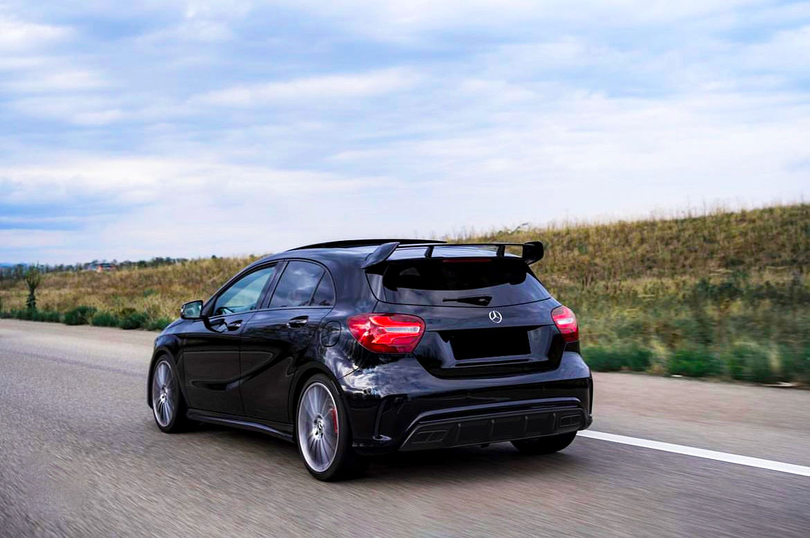 A black car is driving on an open highway, flanked by grassy fields, under a cloudy sky.