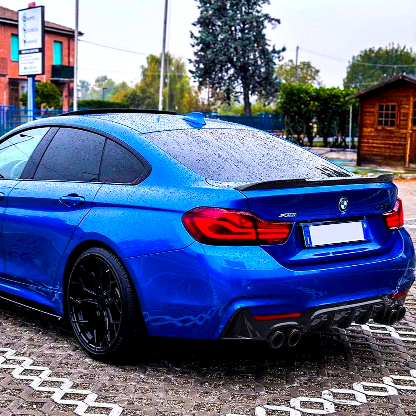 Blue car, parked on wet brick pavement, displaying "BMW" logo and "X Drive 430i" emblem; surrounded by trees, a wooden building, and a sign with illegible text.