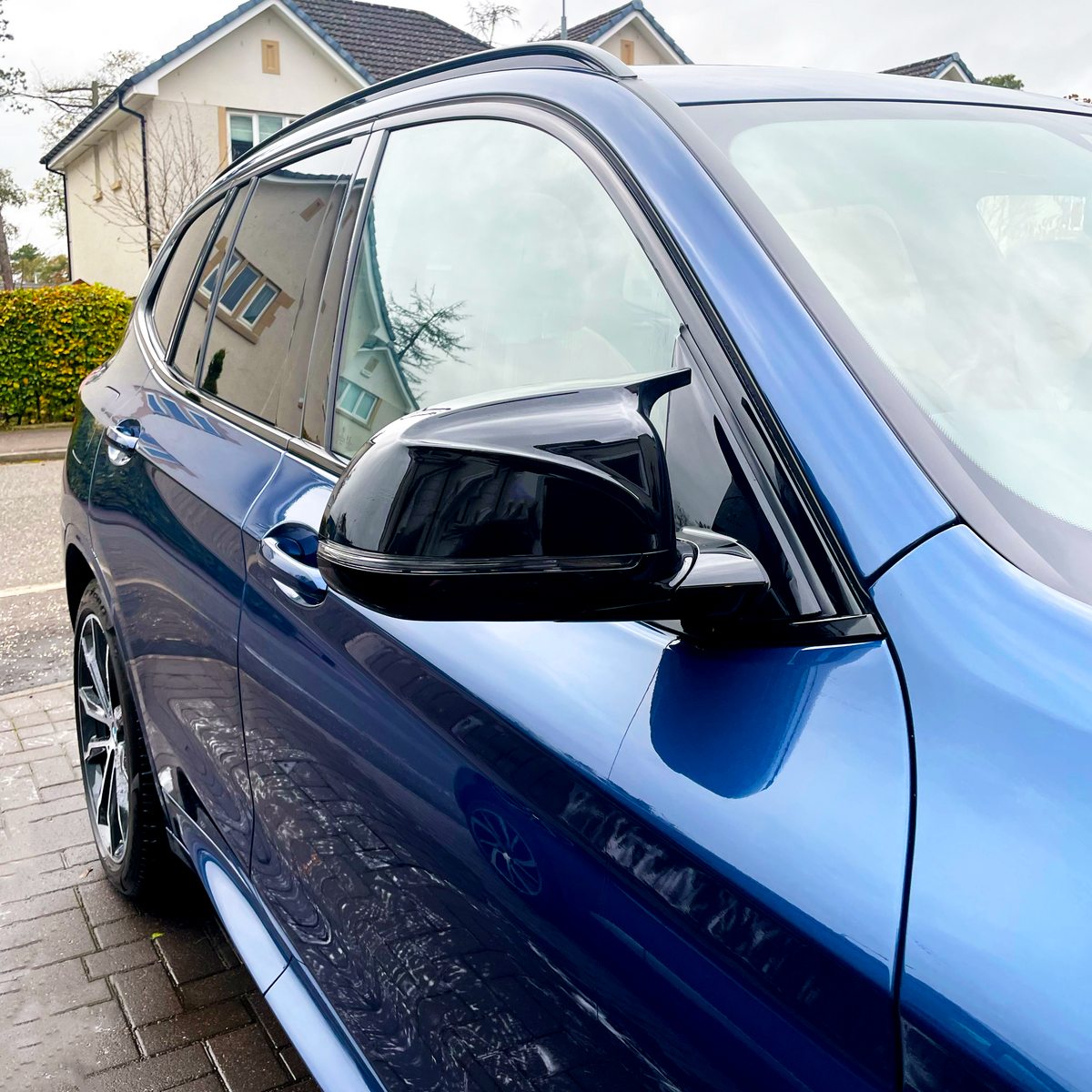 A blue car is parked on a paved driveway with houses and bushes in the background. The car's side mirror and window are prominently visible, reflecting the surroundings.