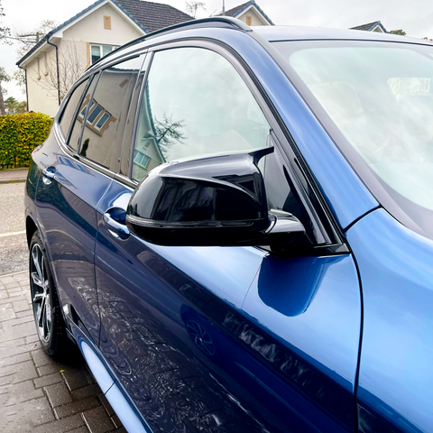 A blue car is parked on a paved driveway with houses and bushes in the background. The car's side mirror and window are prominently visible, reflecting the surroundings.