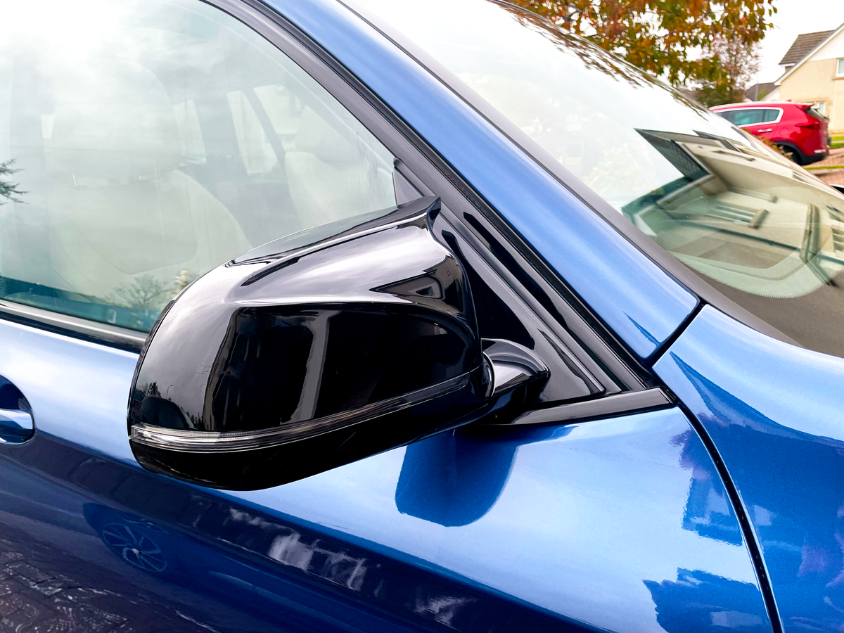 A shiny black side mirror is attached to a blue car, reflecting nearby houses. The car is parked outdoors, with other vehicles visible in the background.