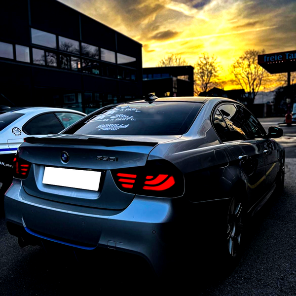 A silver BMW 335i with illuminated rear lights is parked near other vehicles at sunset. The rear window displays the text: "Good life, I say... drive like a maniac... Gorilla Gang."