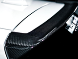 A close-up of a carbon fiber car spoiler on a white vehicle, positioned in a dimly lit garage, with a blurred object and partially visible text reading 