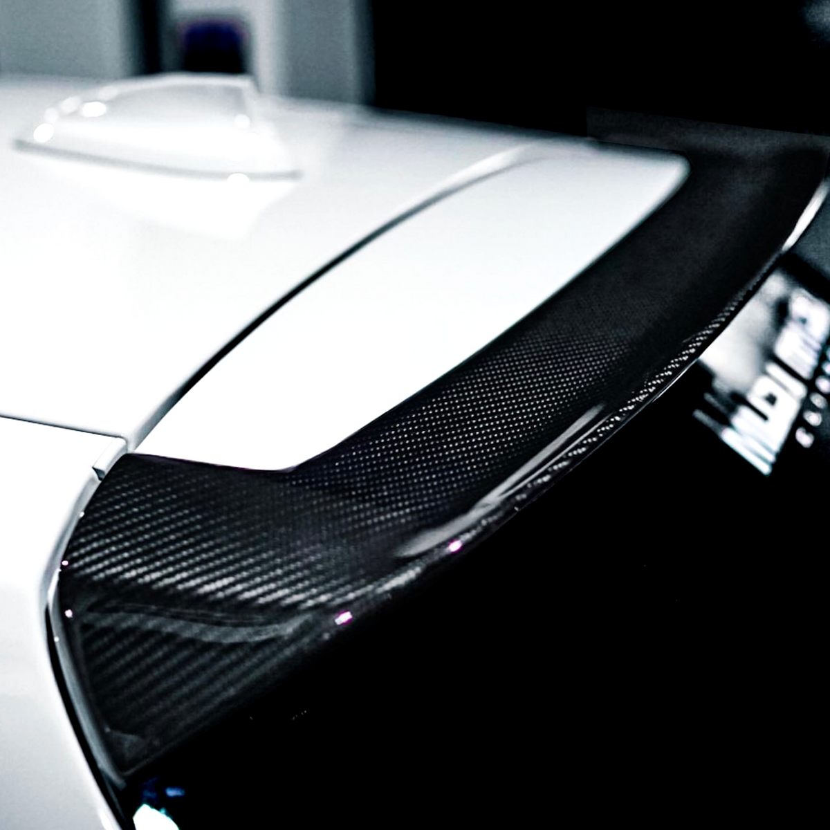 A close-up of a carbon fiber car spoiler on a white vehicle, positioned in a dimly lit garage, with a blurred object and partially visible text reading "MB" in the background.