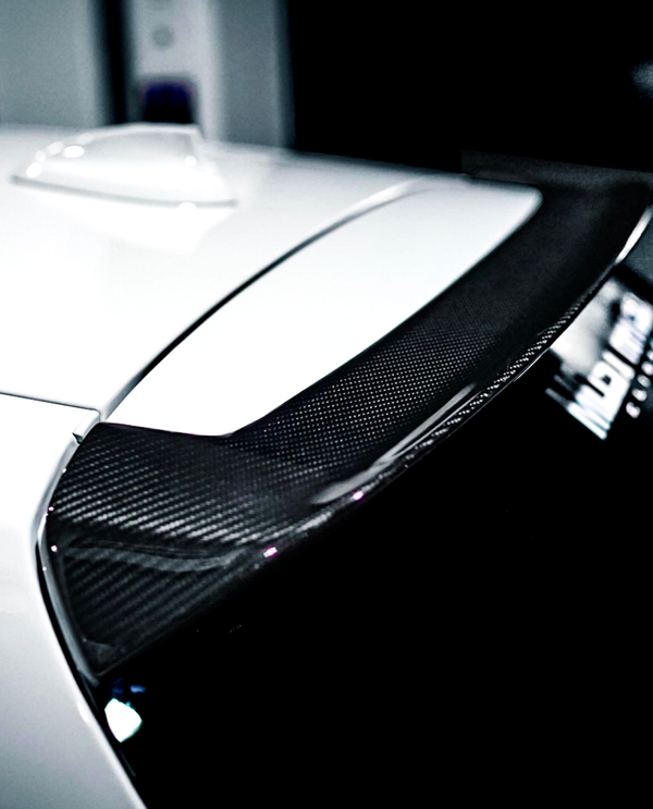 A close-up of a carbon fiber car spoiler on a white vehicle, positioned in a dimly lit garage, with a blurred object and partially visible text reading "MB" in the background.