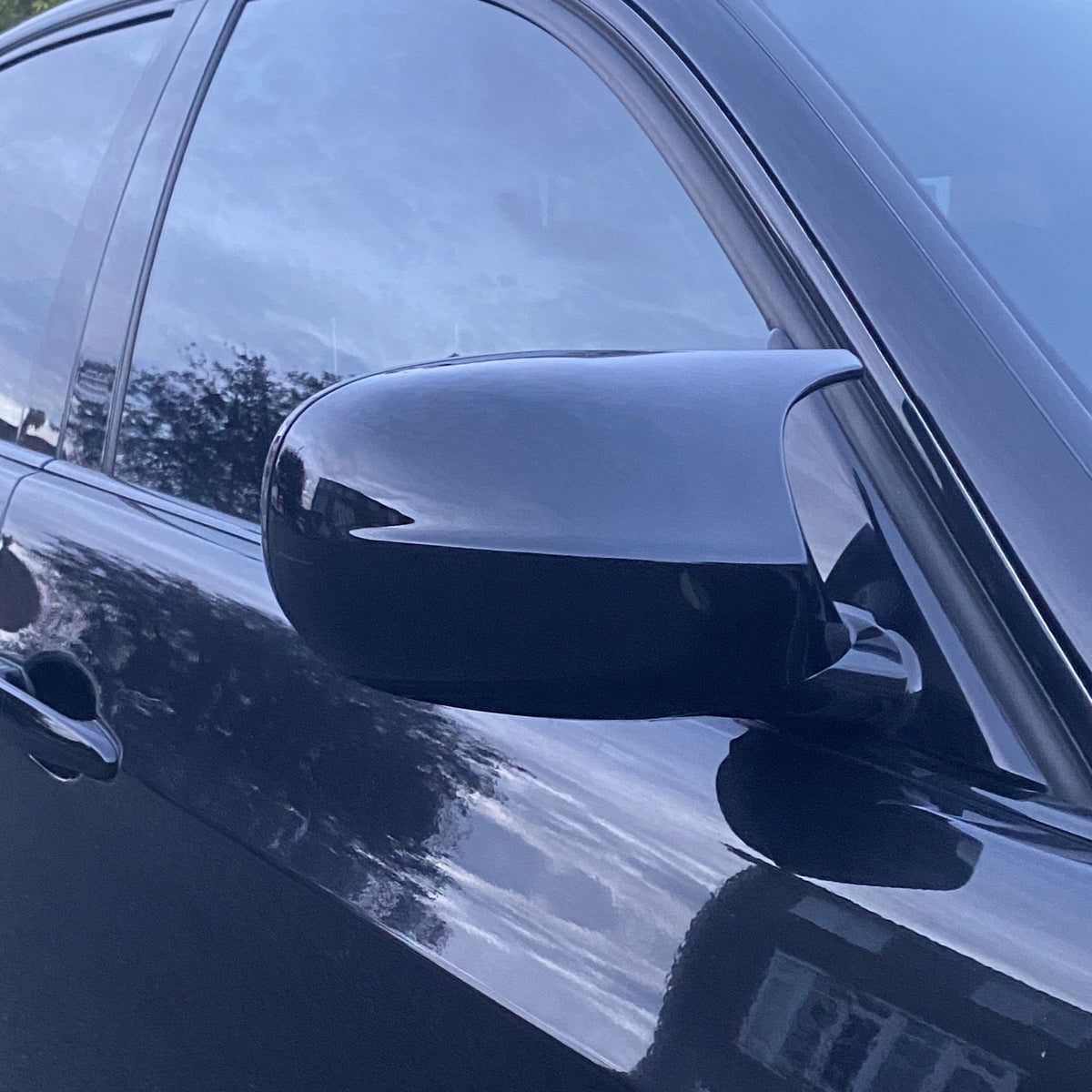 A sleek black car side mirror reflects the evening sky, with blurred greenery and a house in the background, on a parked vehicle.