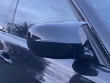 A car's side mirror reflects the surroundings, attached to a sleek, dark vehicle door. The background shows trees and sky, reflecting on the tinted windows and shiny surface.