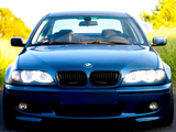 A blue BMW car is parked on a road, surrounded by green foliage under a clear sky. The headlights are on, facing forward.