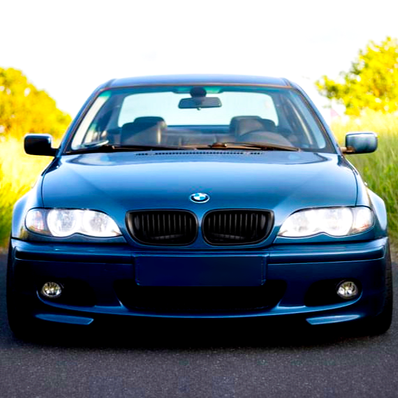 A blue BMW car is parked on a road, surrounded by green foliage under a clear sky. The headlights are on, facing forward.
