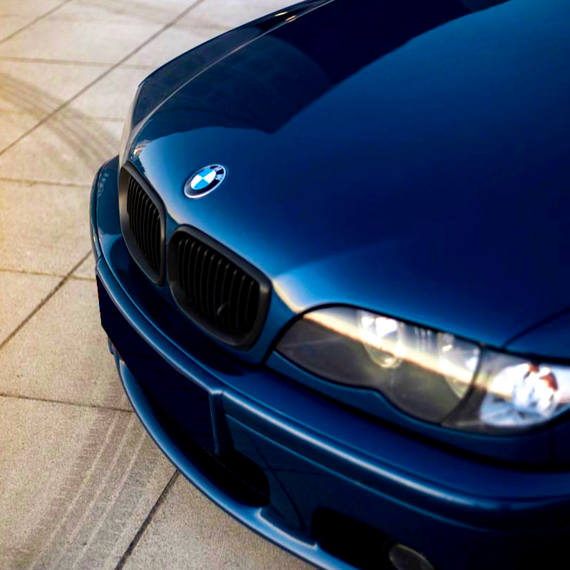 A blue BMW car is parked on a tiled surface, with the focus on its front bumper and headlights, reflecting natural light.