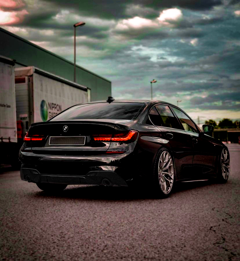 A black car with illuminated rear lights is parked on an empty street. Large wheels and a sleek design stand out. Nearby are cargo trailers under a cloudy sky.