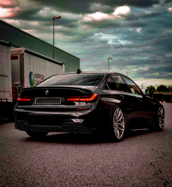 A black car with illuminated rear lights is parked on an empty street. Large wheels and a sleek design stand out. Nearby are cargo trailers under a cloudy sky.
