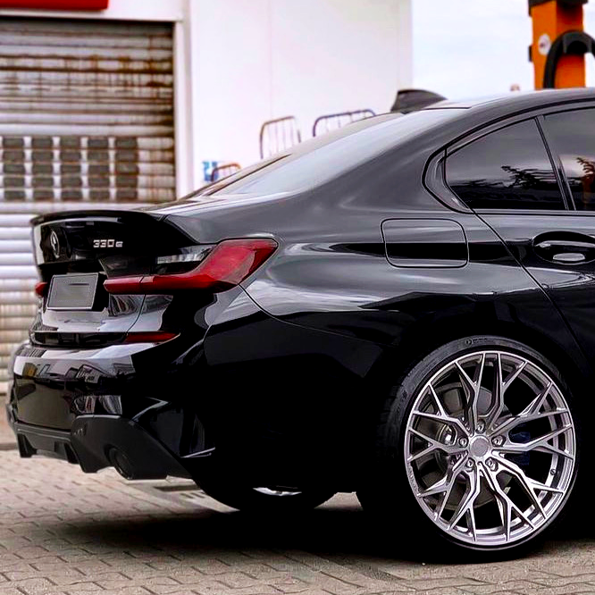 A black BMW 330e parked on a paved surface, showcasing sleek alloy wheels. It is near a shuttered garage in an urban setting.