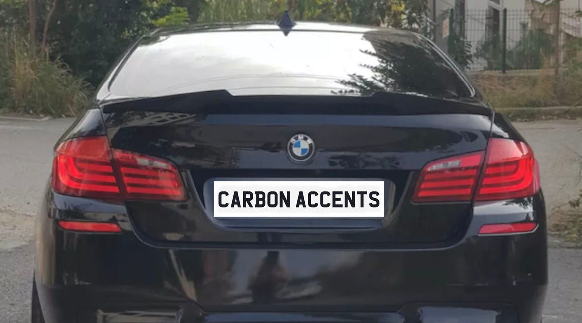 Car parked on a road, showcasing its rear with red taillights, a BMW emblem, and a license plate reading "CARBON ACCENTS." Vegetation lines the roadside.