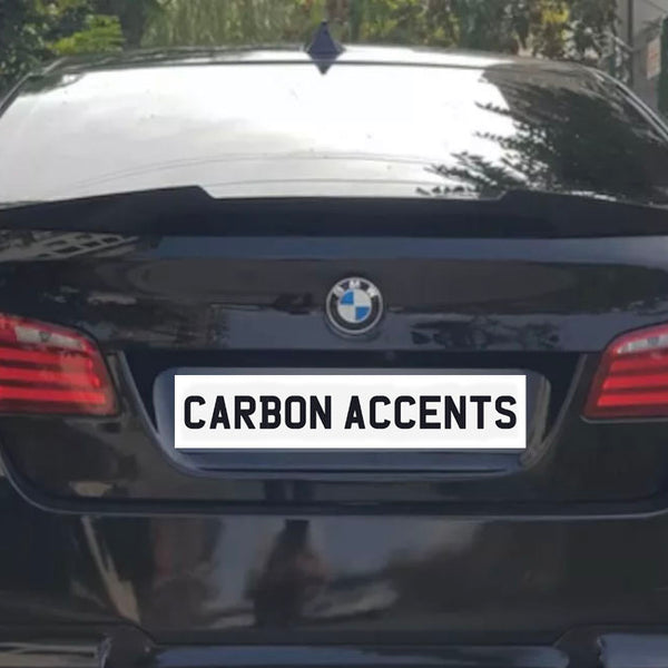 Car parked on a road, showcasing its rear with red taillights, a BMW emblem, and a license plate reading "CARBON ACCENTS." Vegetation lines the roadside.