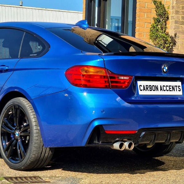 A blue BMW car is parked, showcasing its rear design and dual exhausts. The license plate reads "CARBON ACCENTS" in a sunny, urban setting with brick and glass structures.