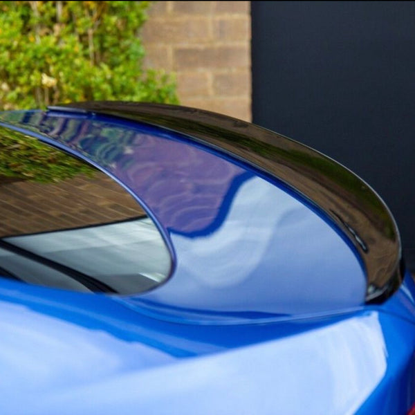 A blue car's rear spoiler reflects its surroundings, parked beside a brick wall and greenery, with a large, dark door in the background providing contrast.