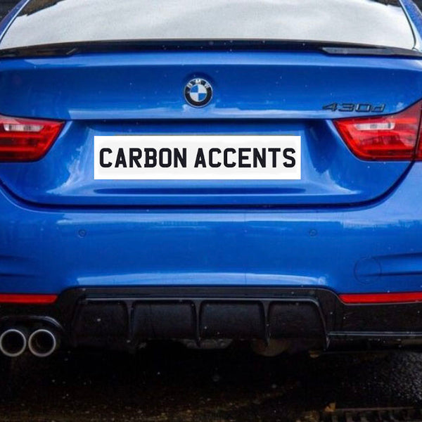 A blue BMW 430d, viewed from the rear, is parked on a wet pavement. The license plate reads "CARBON ACCENTS." Brick walls make up the background.