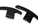 Two carbon-fiber paddle shifters, positioned side-by-side, lie on a plain white background. The right paddle features a plus sign and an arrow.