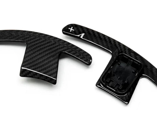 Two carbon-fiber paddle shifters, positioned side-by-side, lie on a plain white background. The right paddle features a plus sign and an arrow.