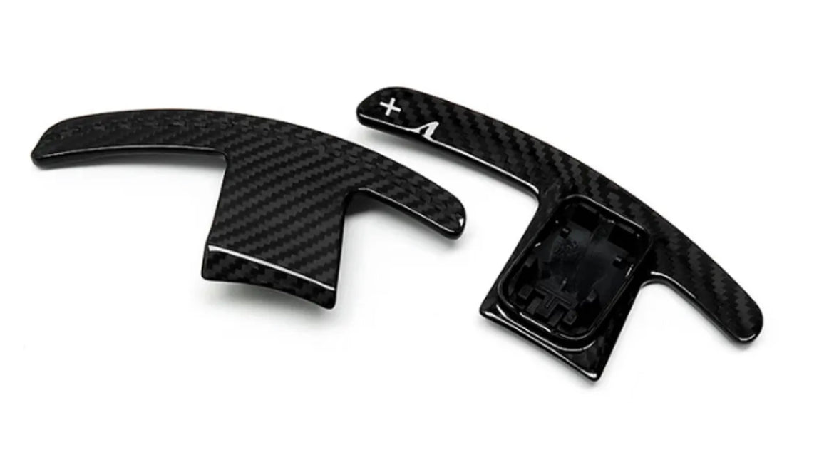 Two carbon-fiber paddle shifters, positioned side-by-side, lie on a plain white background. The right paddle features a plus sign and an arrow.