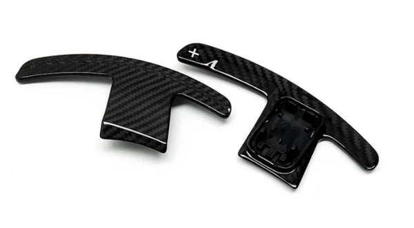 Two carbon-fiber paddle shifters, positioned side-by-side, lie on a plain white background. The right paddle features a plus sign and an arrow.