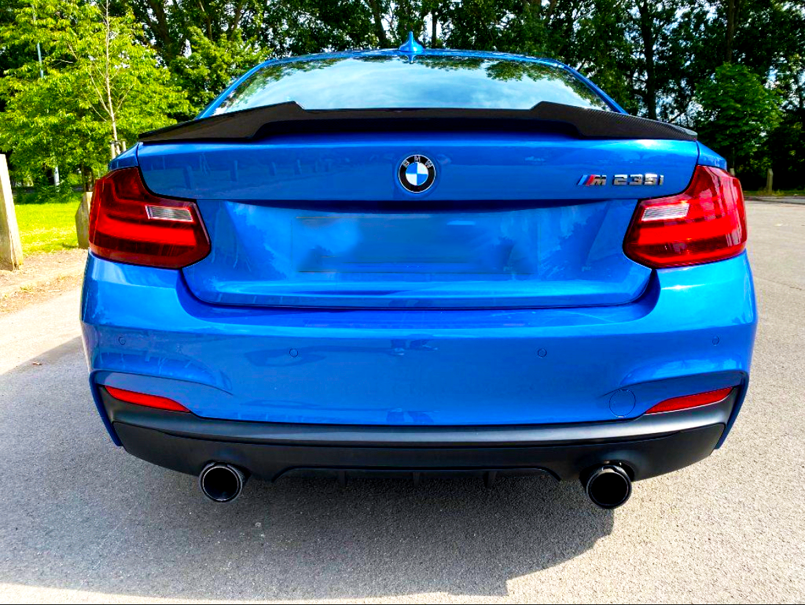 A blue BMW M235i is parked on the side of a road. The sleek car is surrounded by lush green trees.