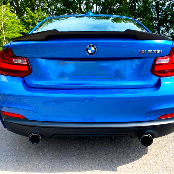 A blue BMW M235i is parked on the side of a road. The sleek car is surrounded by lush green trees.
